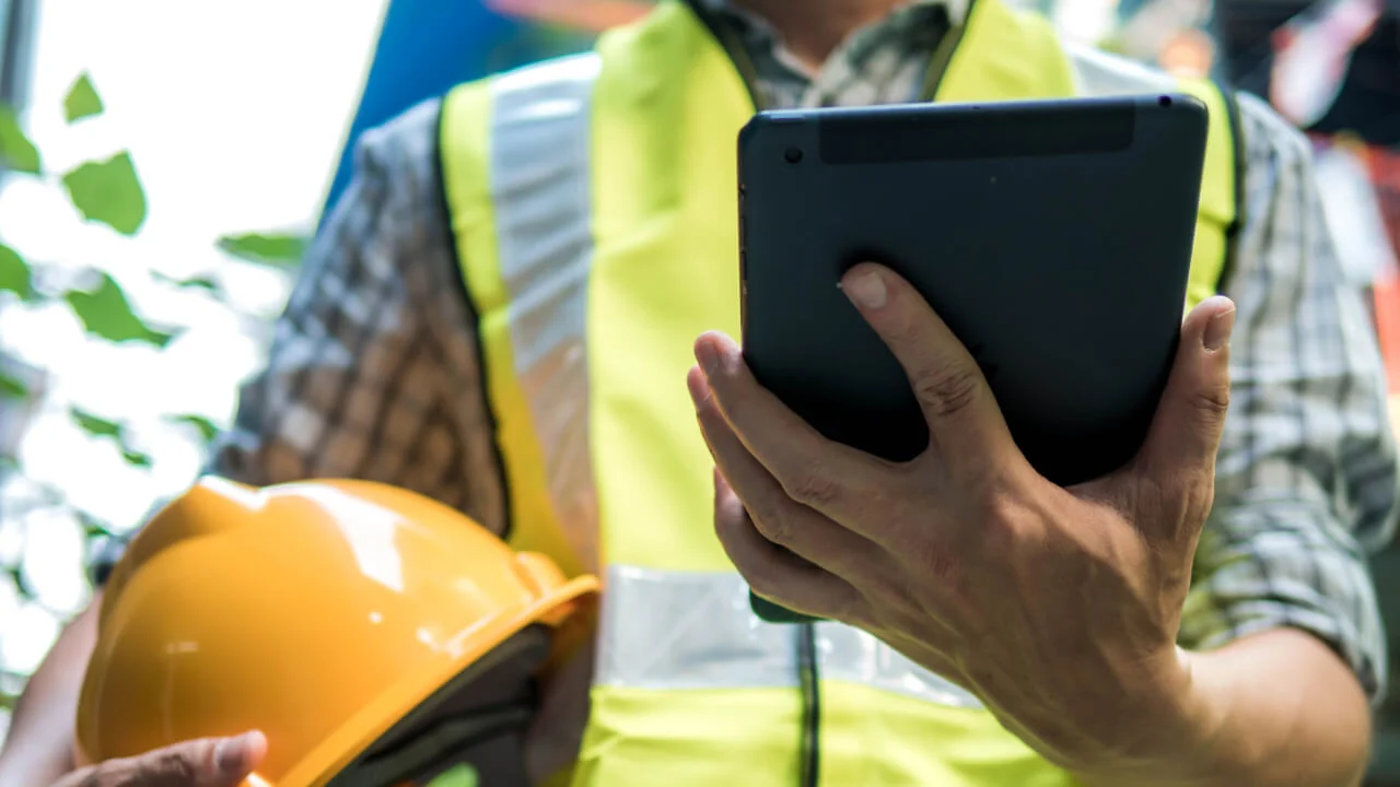 Worker in a reflective vest holding a hard hat and tablet